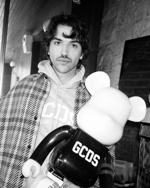 Monochrome portrait of a man in a plaid jacket holding a bear-shaped toy with branding in an indoor setting with brick walls.