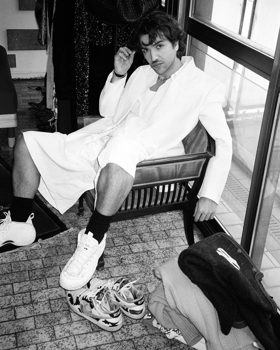 Monochrome portrait of a man with a mustache sitting indoors in a suit jacket, shorts, and sneakers, with shoes on the floor.