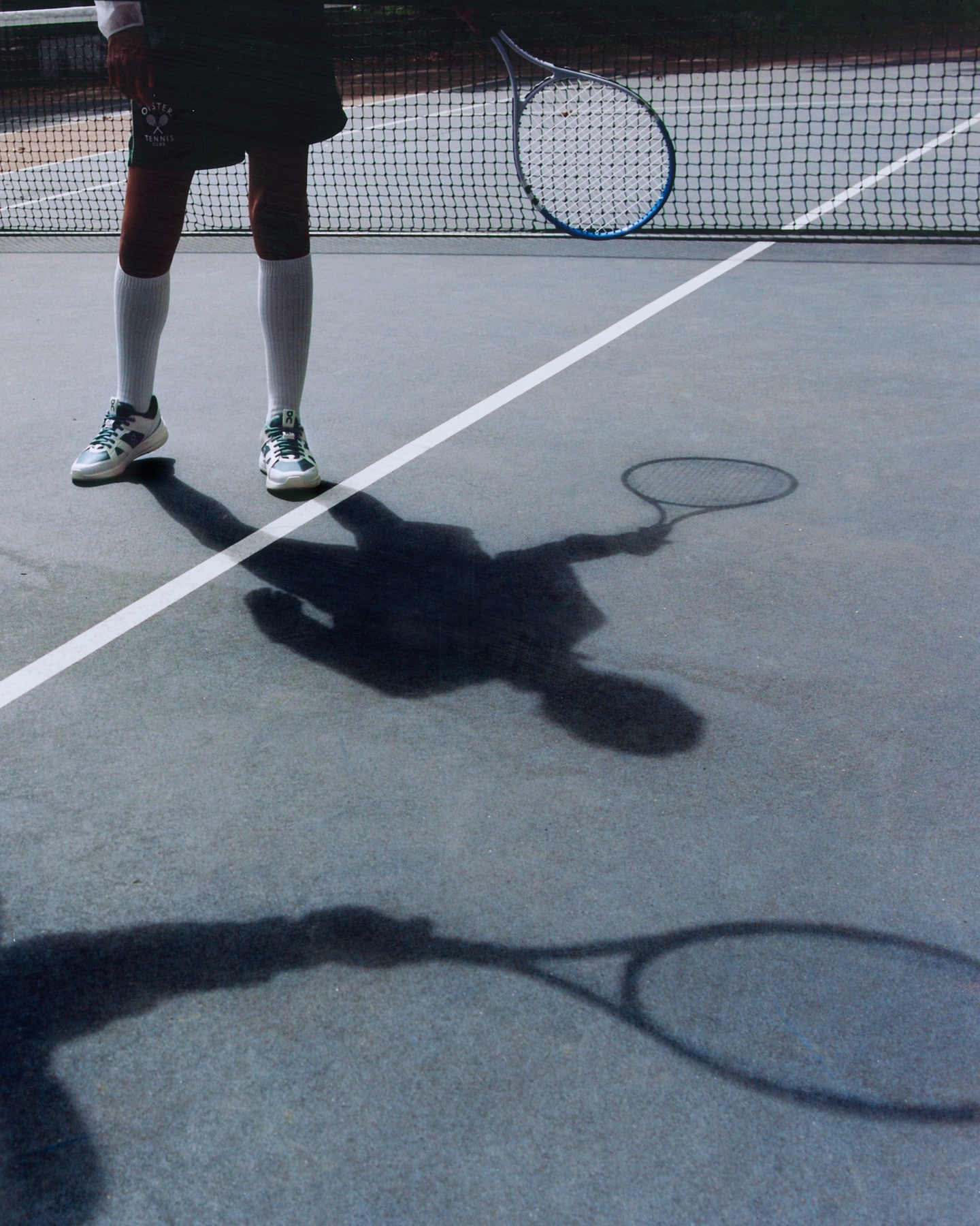 Tennis player stands on a grey outdoor court with a tennis racket; the tennis net is visible in the background, and the player's shadow is cast on the court.