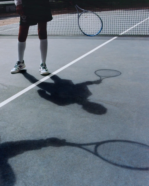 Tennis player stands on a grey outdoor court with a tennis racket; the tennis net is visible in the background, and the player's shadow is cast on the court.
