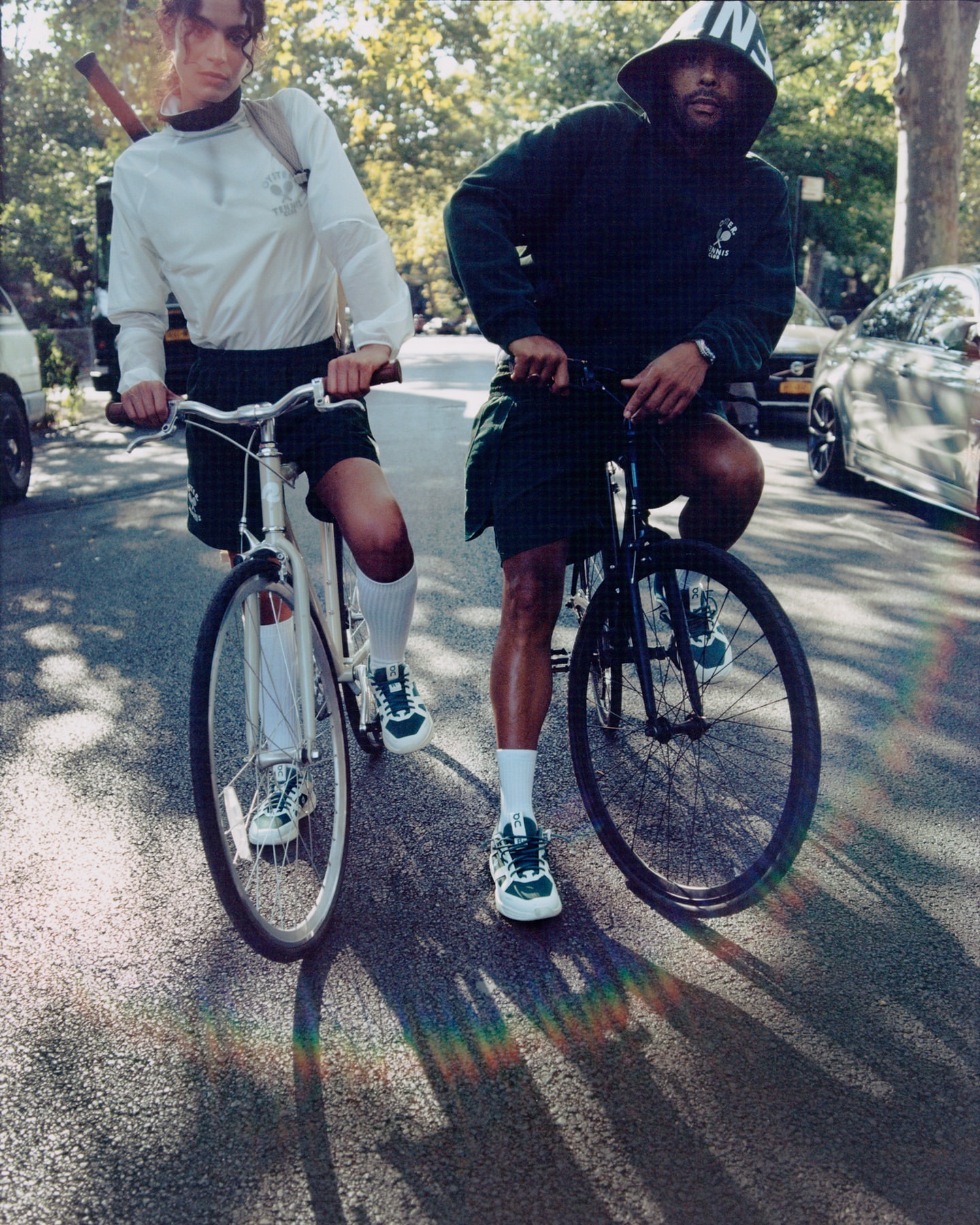 Two adult males on bicycles in a street setting with cars and trees visible in the background, shot in daylight, styled for a casual look.