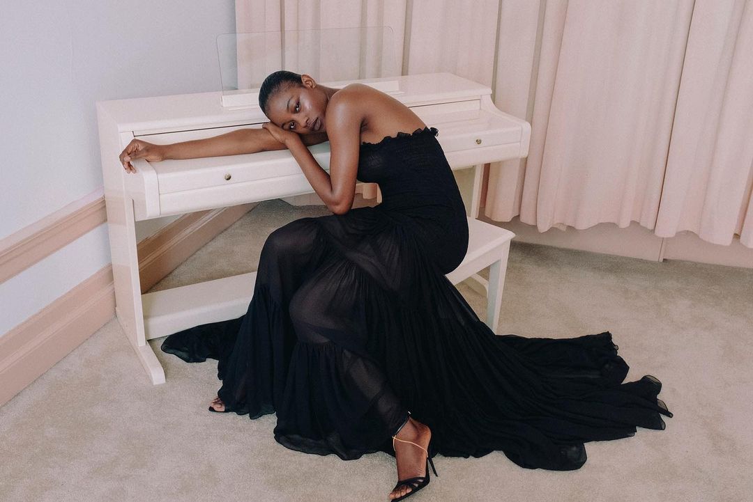 Elegant African-American woman in a black gown poses gracefully beside a white piano in a minimalist studio setting. Fashion editorial.