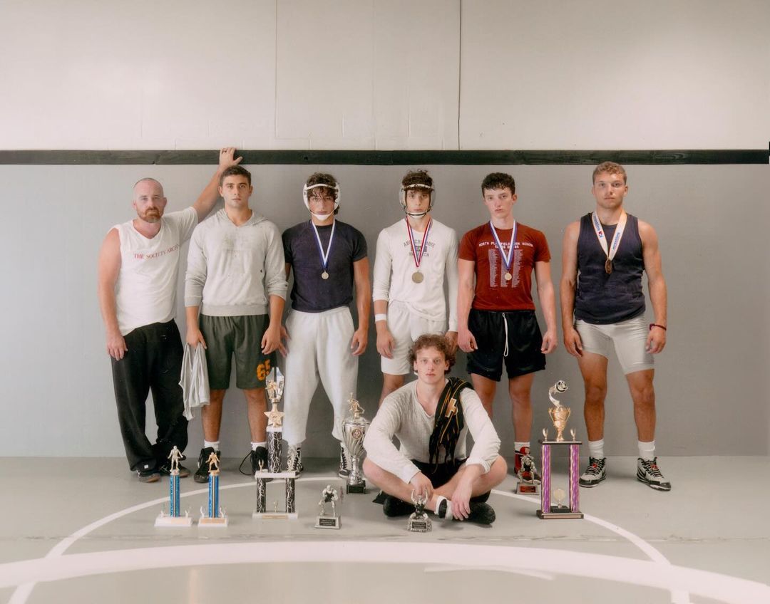 Seven male athletes in wrestling attire and medals stand near trophies on a wrestling mat against a plain wall backdrop in a studio.