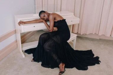 Black woman in a long black dress reclining against a white upright piano in a room with light carpet and pleated curtains.