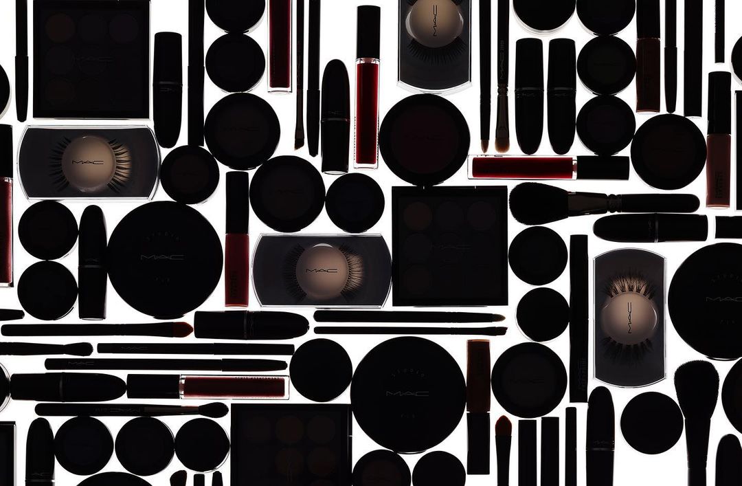 High-angle, full-frame product photography of a variety of MAC makeup products in black packaging on a white background, creating a seamless pattern.
