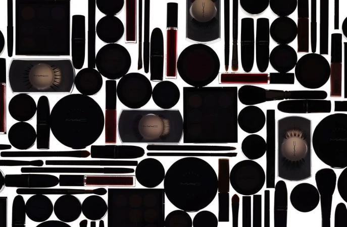 High-angle, full-frame product photography of a variety of MAC makeup products in black packaging on a white background, creating a seamless pattern.