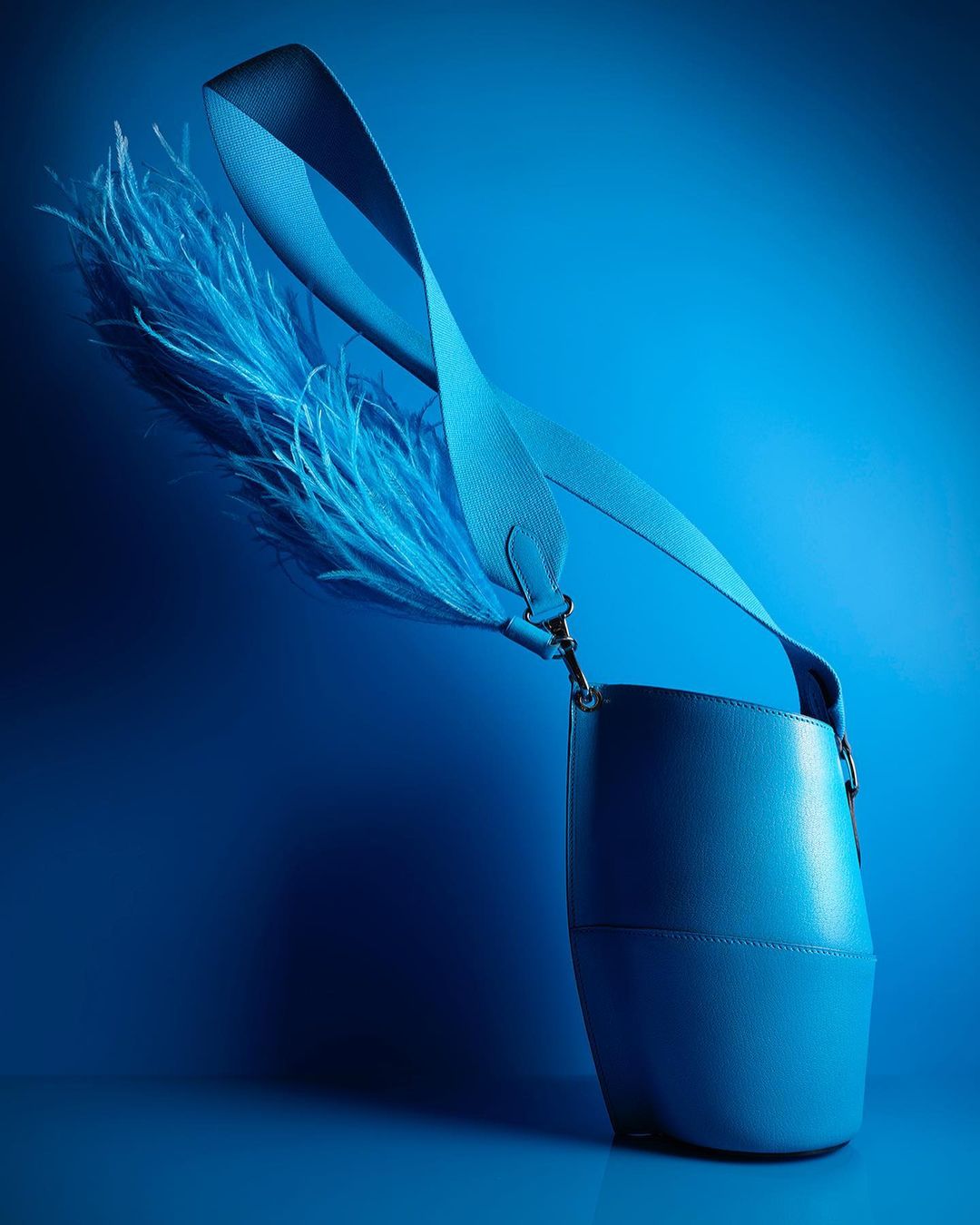 Elegant light blue leather bucket bag with a wide strap and feathered tassels, set against a graduated blue background.