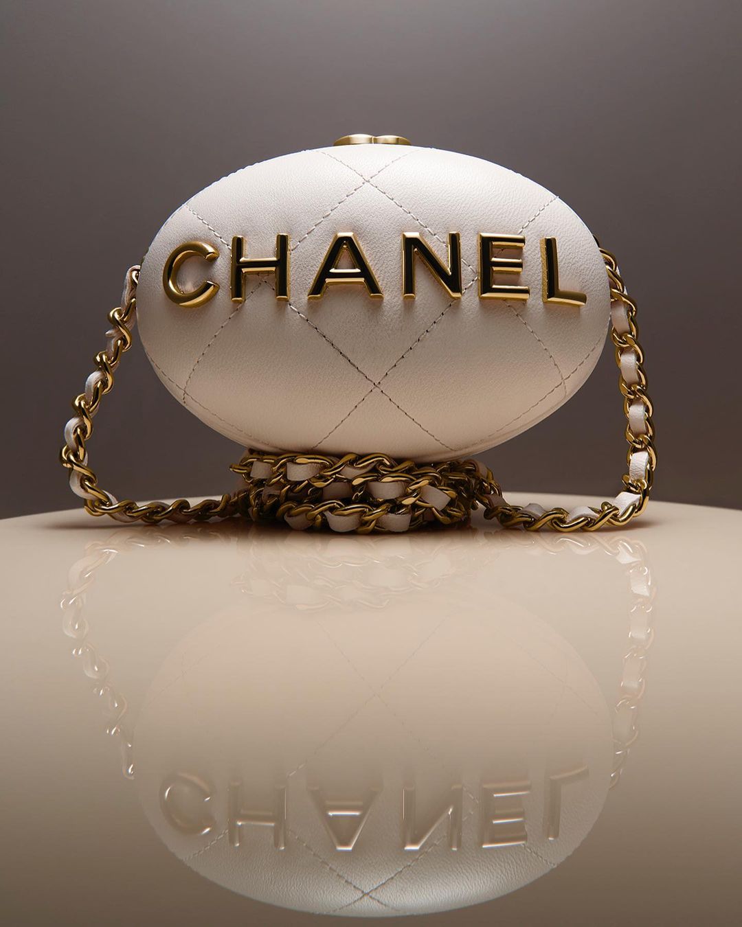 Elegant Chanel egg minaudiere bag with golden chain strap and white quilted leather, displayed on a glossy surface with reflection.