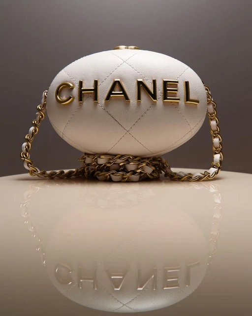 Elegant Chanel egg minaudiere bag with golden chain strap and white quilted leather, displayed on a glossy surface with reflection.