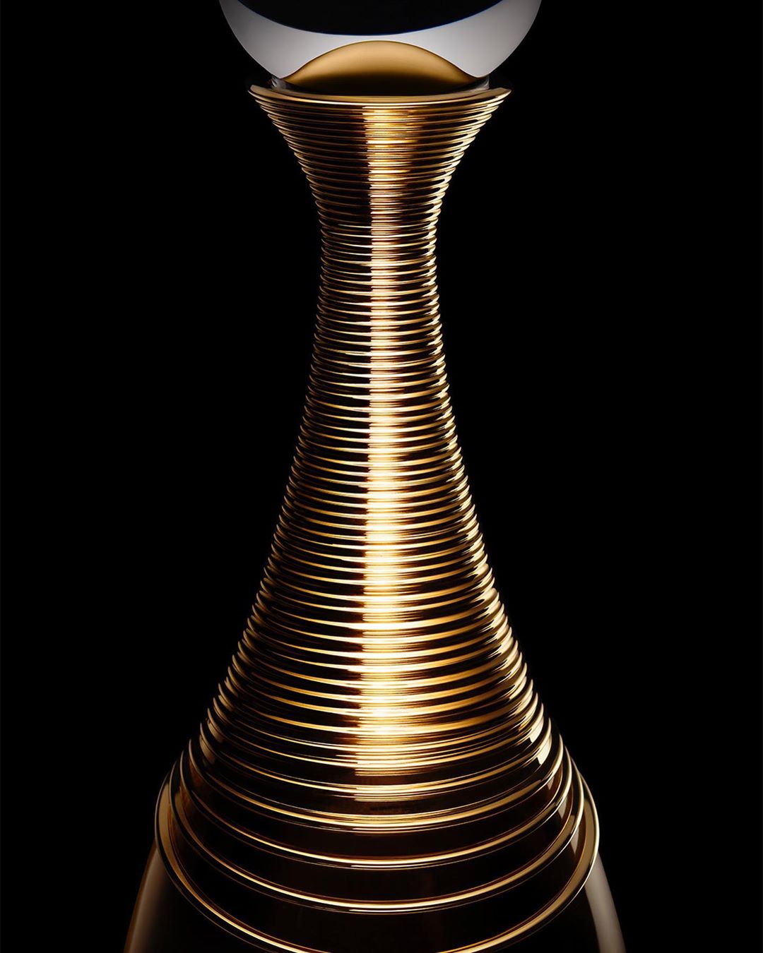 Elegant gold perfume bottle with sleek, ribbed design, white orb top, set against a stark black background in a studio-lit product shot.
