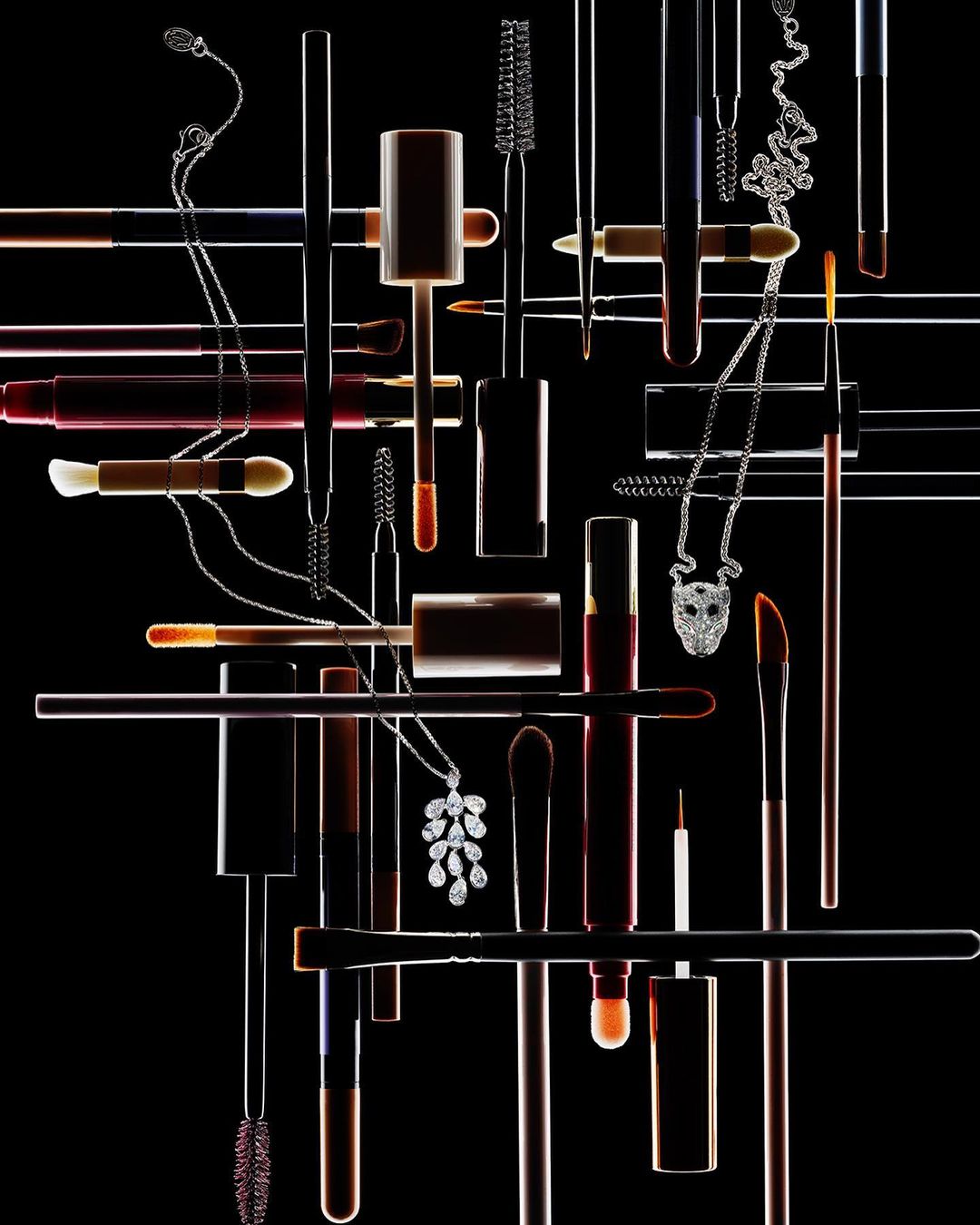 Still life shot of multiple makeup brushes, lipstick tubes, and necklaces against a black backdrop, with contrasting lighting.
