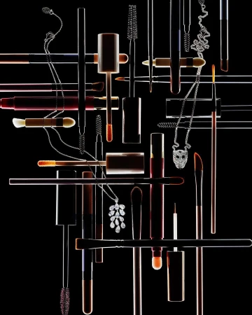 Still life shot of multiple makeup brushes, lipstick tubes, and necklaces against a black backdrop, with contrasting lighting.