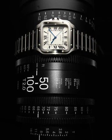 Luxury silver Cartier Santos wristwatch on a camera lens, studio shot. High-end timekeeping, focus on precision watchmaking craftsmanship.