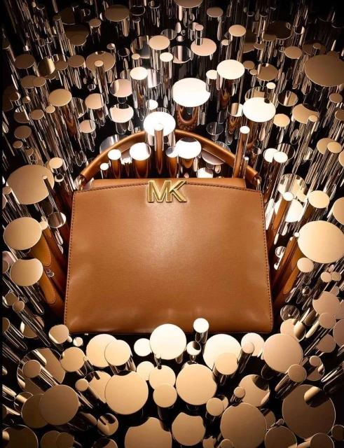A tan Michael Kors handbag is showcased against a backdrop of reflective gold and silver cylindrical tubes and circles, with the MK logo.