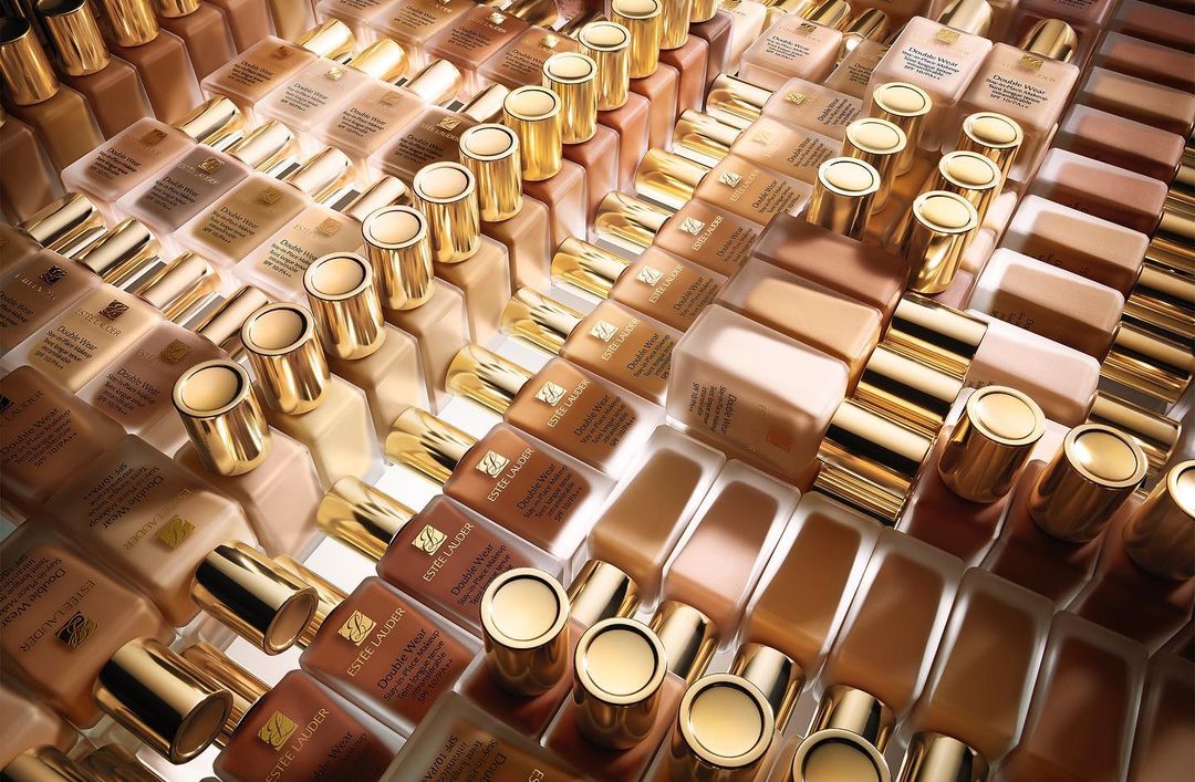 High-angle, overhead shot of many bottles of Estée Lauder Double Wear foundation in various shades, neatly arranged on a surface.