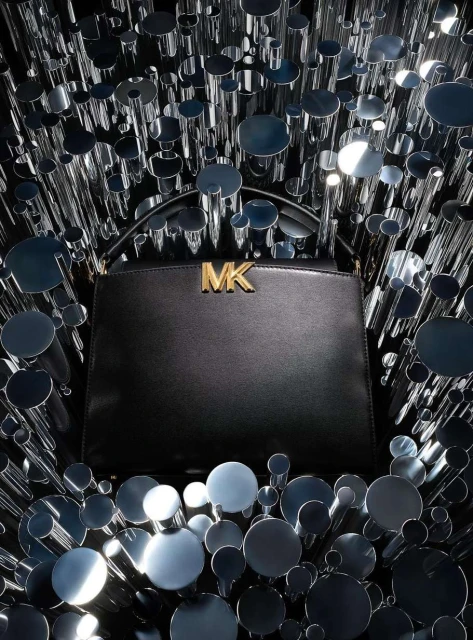 Michael Kors handbag in black, featuring gold logo hardware. Cylindrical reflective elements add a modern, abstract touch.