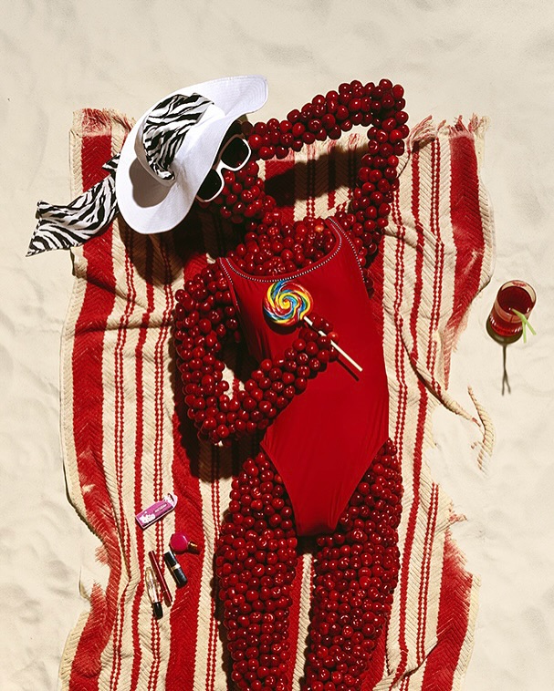 Creative beach scene: red berries arranged to resemble a woman in a swimsuit with accessories like a hat and lollipop.