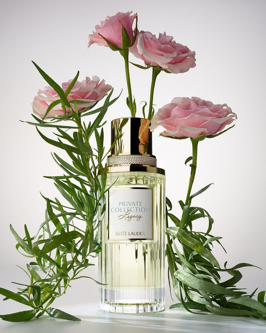 Estee Lauder Private Collection Legacy perfume bottle surrounded by pink roses and green foliage against a light background in a still life shot.
