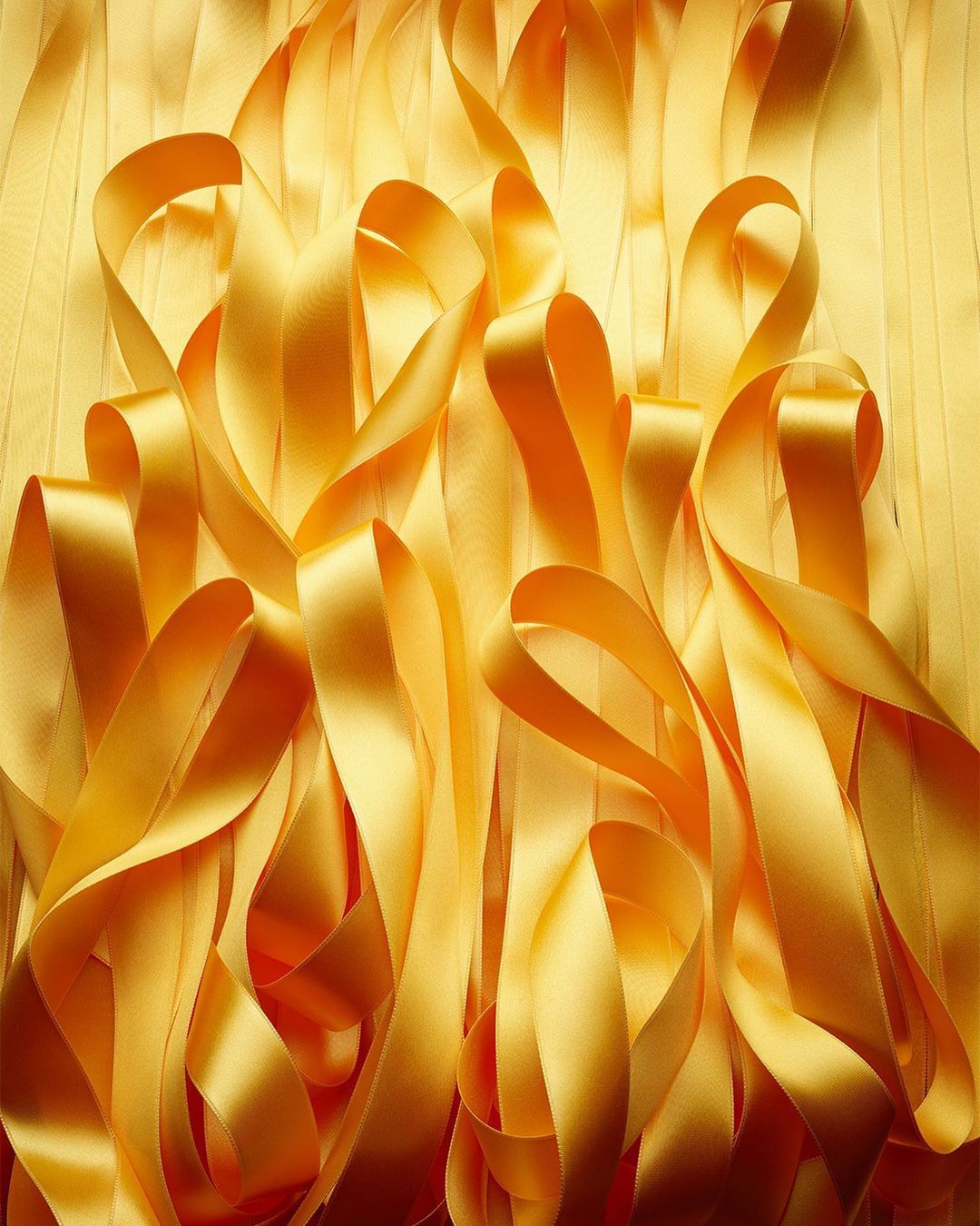 Close-up of many looping strands of golden yellow satin ribbons in a vertical composition with a soft, flowing abstract design.