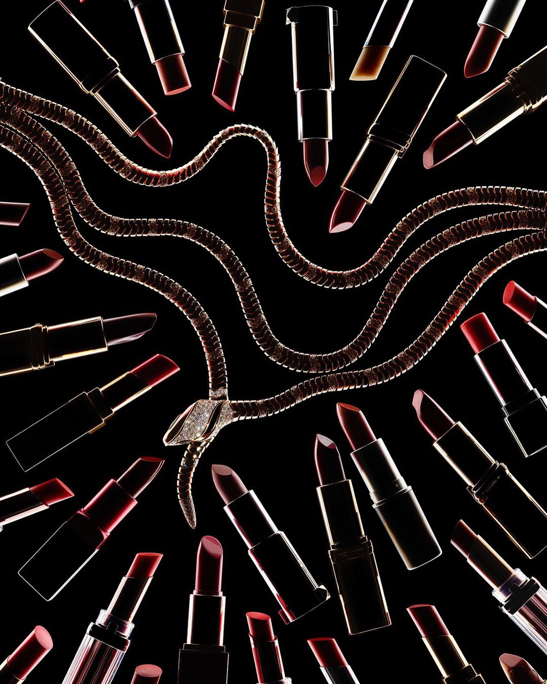 High-angle studio shot of a snake necklace surrounded by an array of dark red lipsticks against a stark black background. Beauty and fashion.