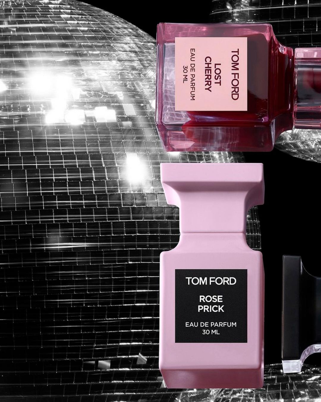 Two Tom Ford perfume bottles, 'Lost Cherry' and 'Rose Prick,' stand against a backdrop of shimmering disco balls and glossy black.