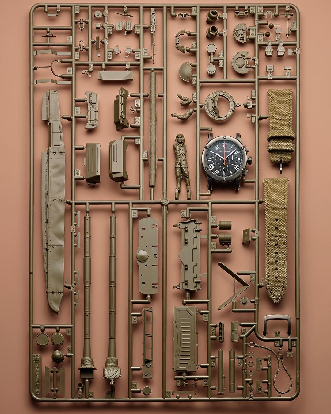 Overhead shot of a watch and plastic model kit parts arranged on a sprue on a pink backdrop, showcasing miniature figures and watch components.