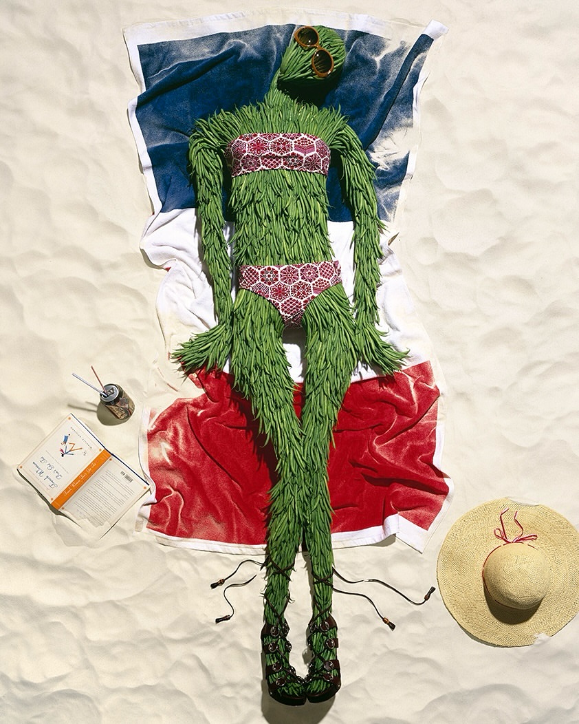 Whimsical image of a green humanoid-like figure sunbathing on a towel in the sand, wearing sunglasses and a bikini, with a beach hat.