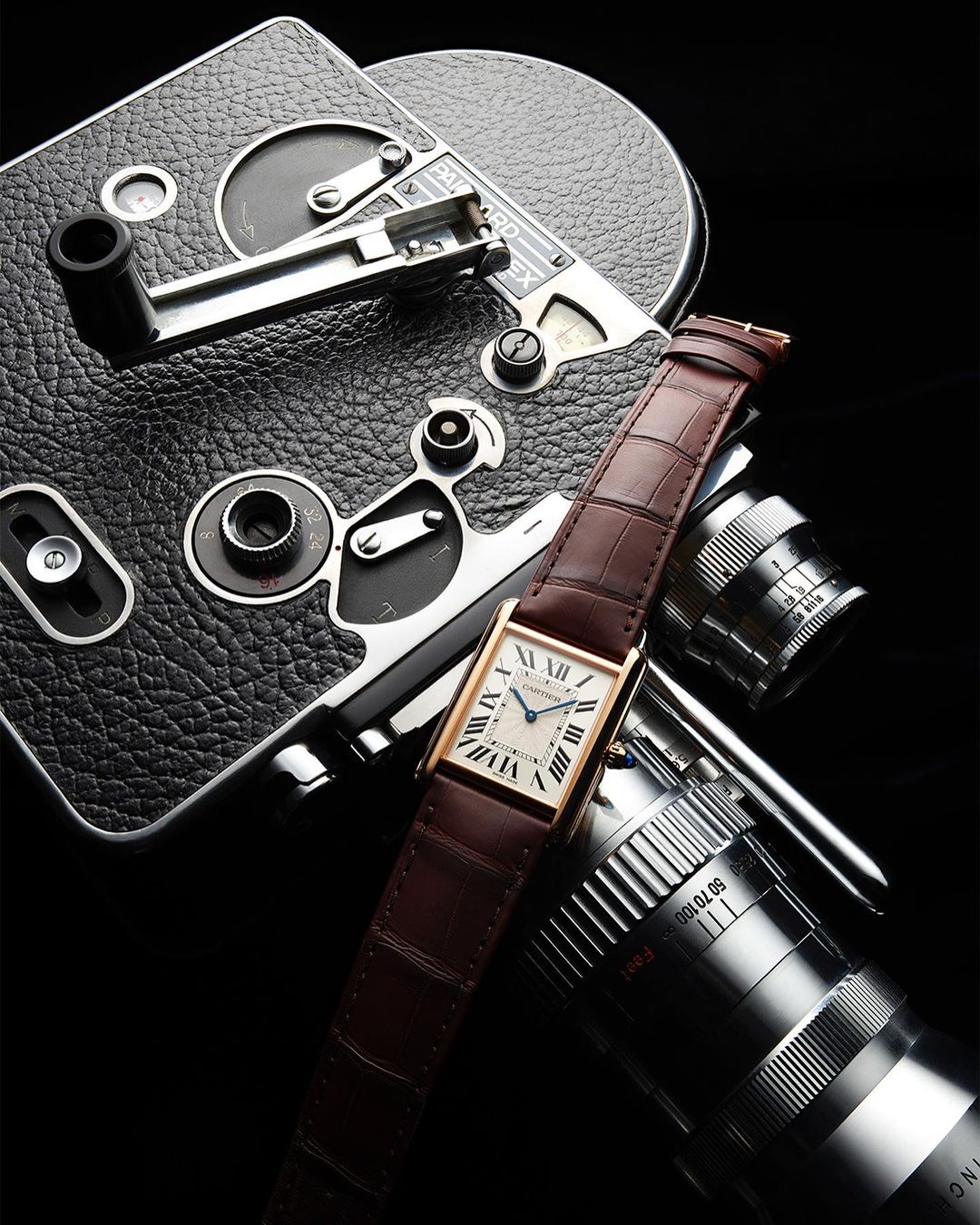Elegant watch with brown leather strap positioned over a vintage movie camera with multiple lenses against a stark black backdrop.
