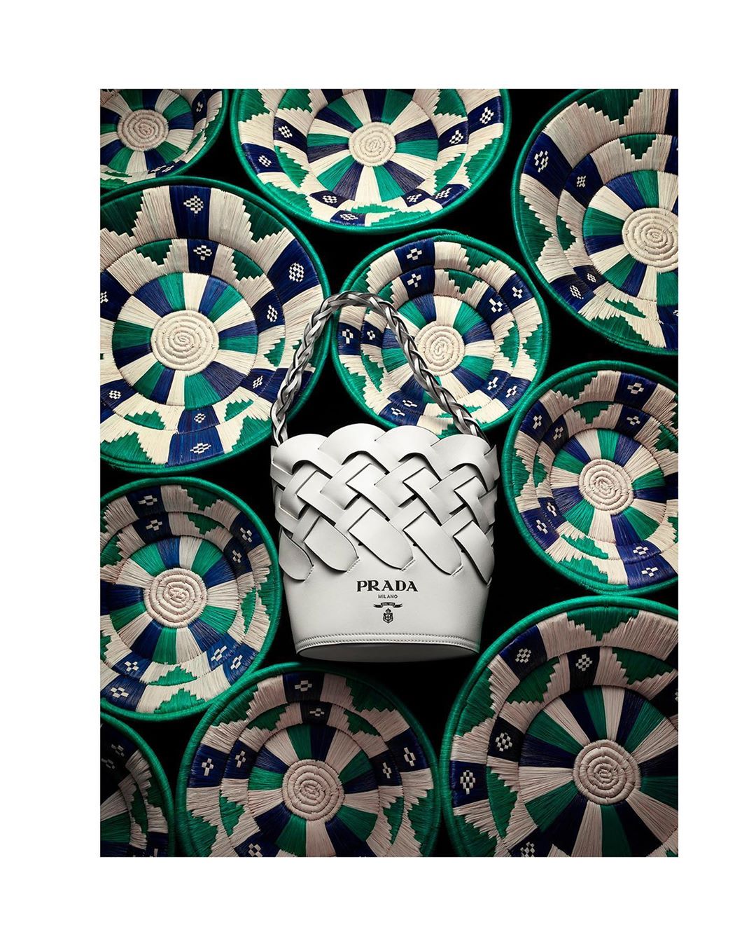 Elevated view of a white woven Prada handbag centered among several green and blue woven straw baskets on a dark background.