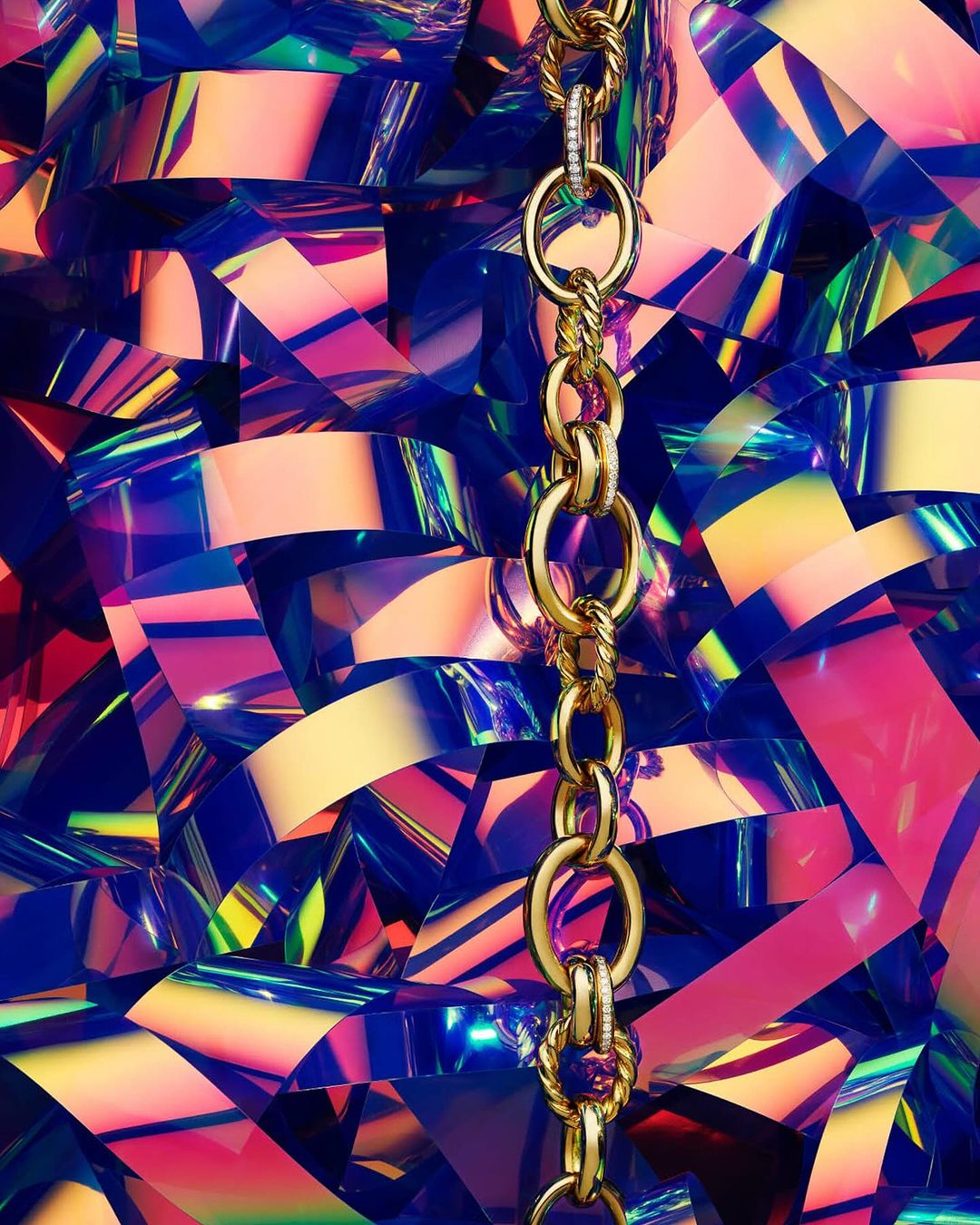 A gold chain necklace is centered over a geometric background of vibrant blue, yellow and pink glossy confetti shapes.