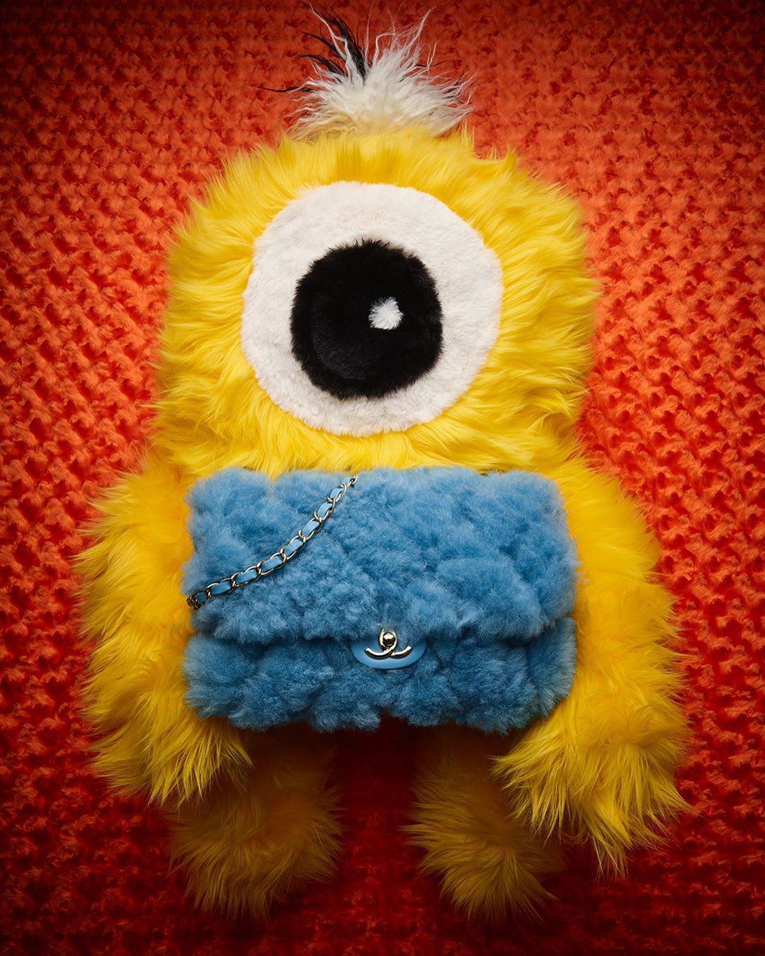 Yellow furry monster-like handbag with a blue quilted flap and chain strap on an orange knitted background, showcasing a whimsical fashion accessory.