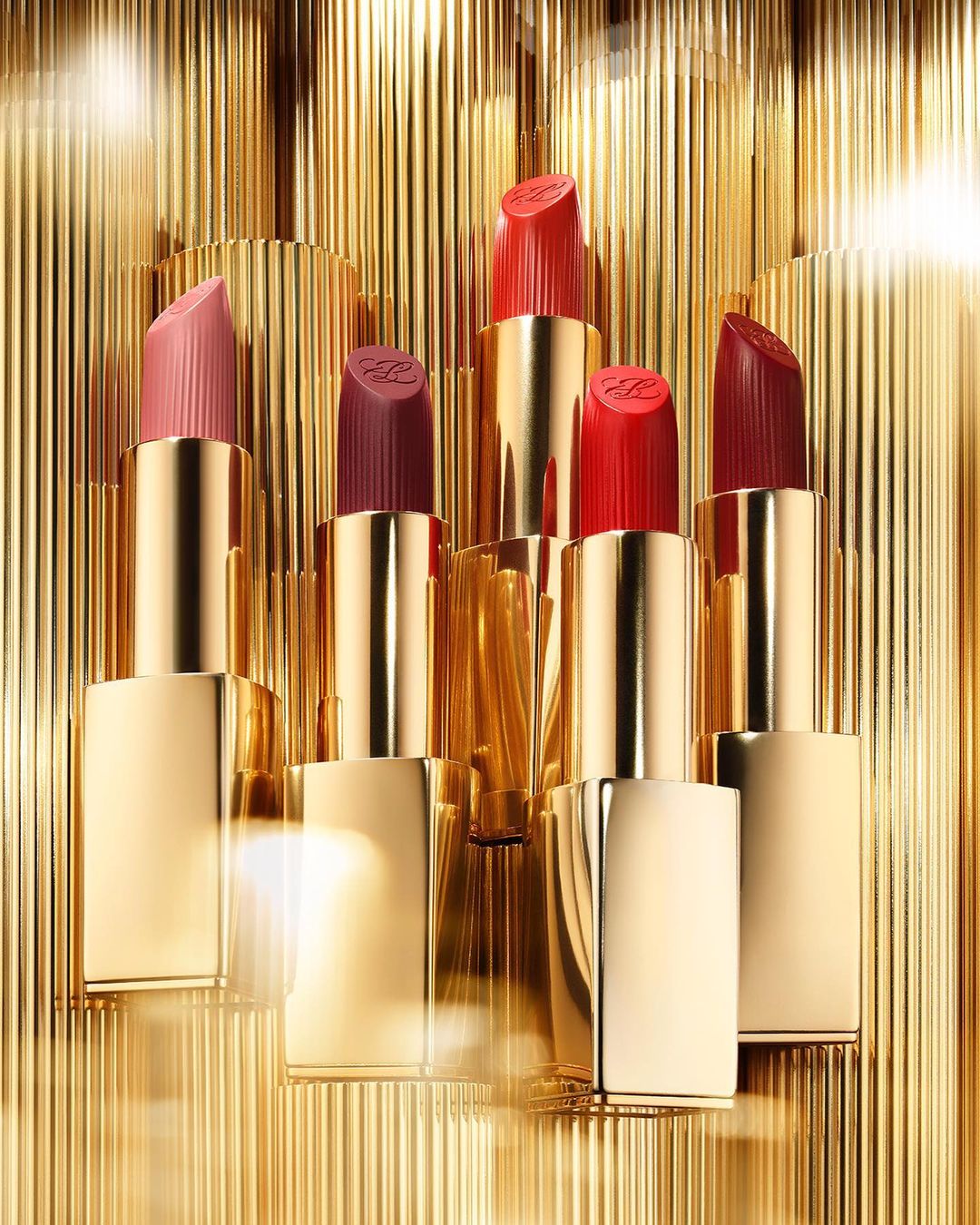 Group of lipsticks in varying shades of red and pink with gold tubes, arranged against a textured gold background.