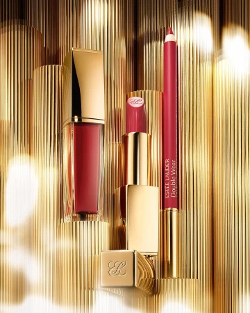 Beauty product still life featuring lipstick, lip gloss, and lip liner by Estee Lauder, against a gold, ridged background.