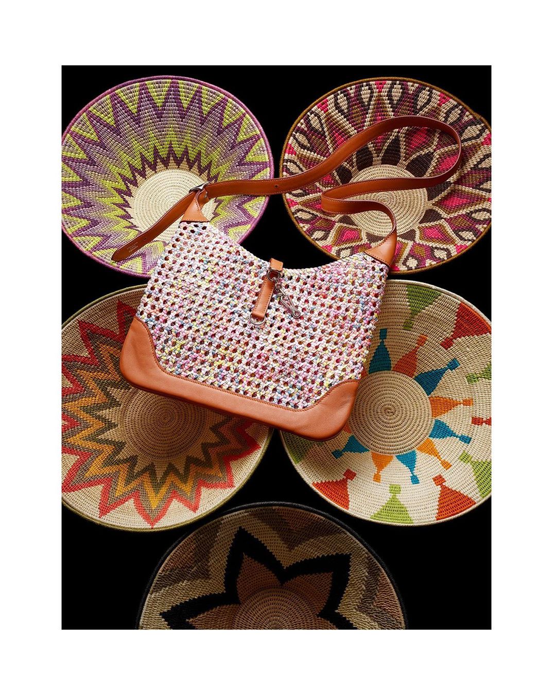 Woven bag with leather trim displayed in front of colorful, circular, woven decorative plates in a studio setting with a black backdrop.