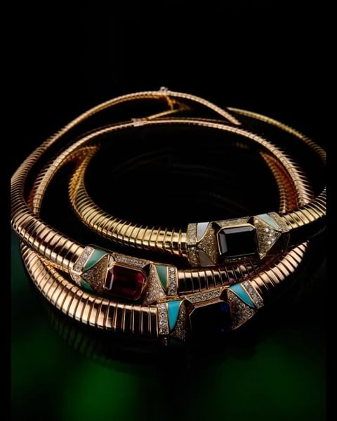Three gold necklaces featuring colored gemstones. The necklaces are coiled against a dark backdrop with a green gradient at the base.