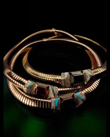 Three gold necklaces featuring colored gemstones. The necklaces are coiled against a dark backdrop with a green gradient at the base.