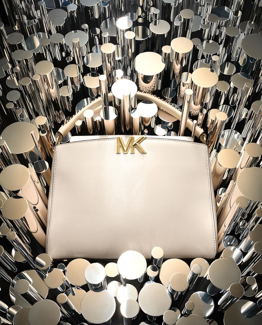 Michael Kors handbag with logo detail. Shiny silver and gold cylinders surround the purse against a black backdrop in a studio setting.