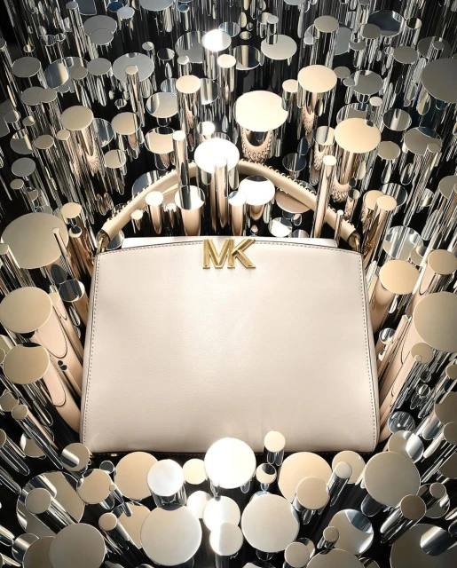 Michael Kors handbag with logo detail. Shiny silver and gold cylinders surround the purse against a black backdrop in a studio setting.