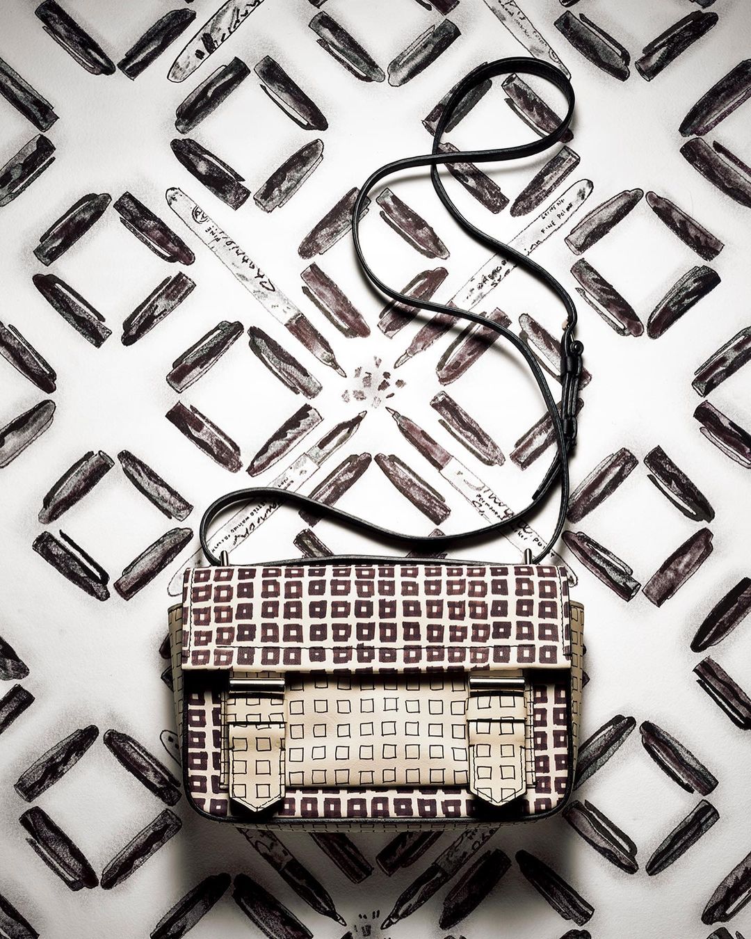 Elevated view of a cream-colored satchel handbag with a grid pattern, black shoulder strap, and a background of markers on white.