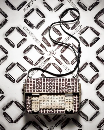 Elevated view of a cream-colored satchel handbag with a grid pattern, black shoulder strap, and a background of markers on white.