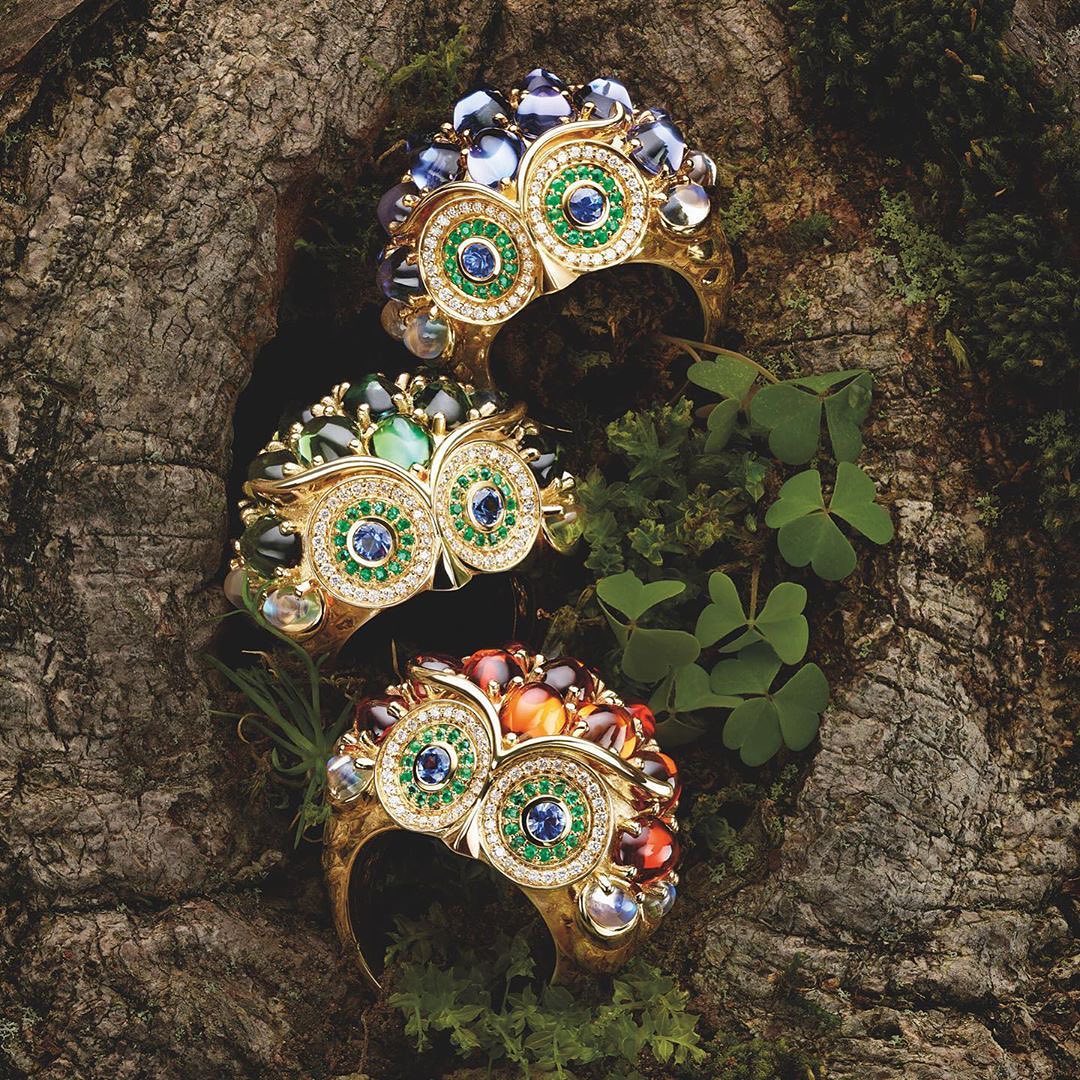 Three gemstone bracelets with a floral owl-shaped design are arranged on a textured tree bark surface with green foliage accents.