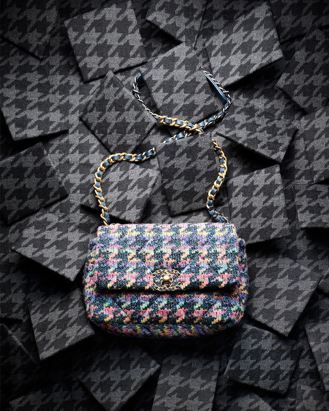 Close-up of a multicolored tweed Chanel flap bag with gold chain strap, laid on gray houndstooth patterned textile squares.