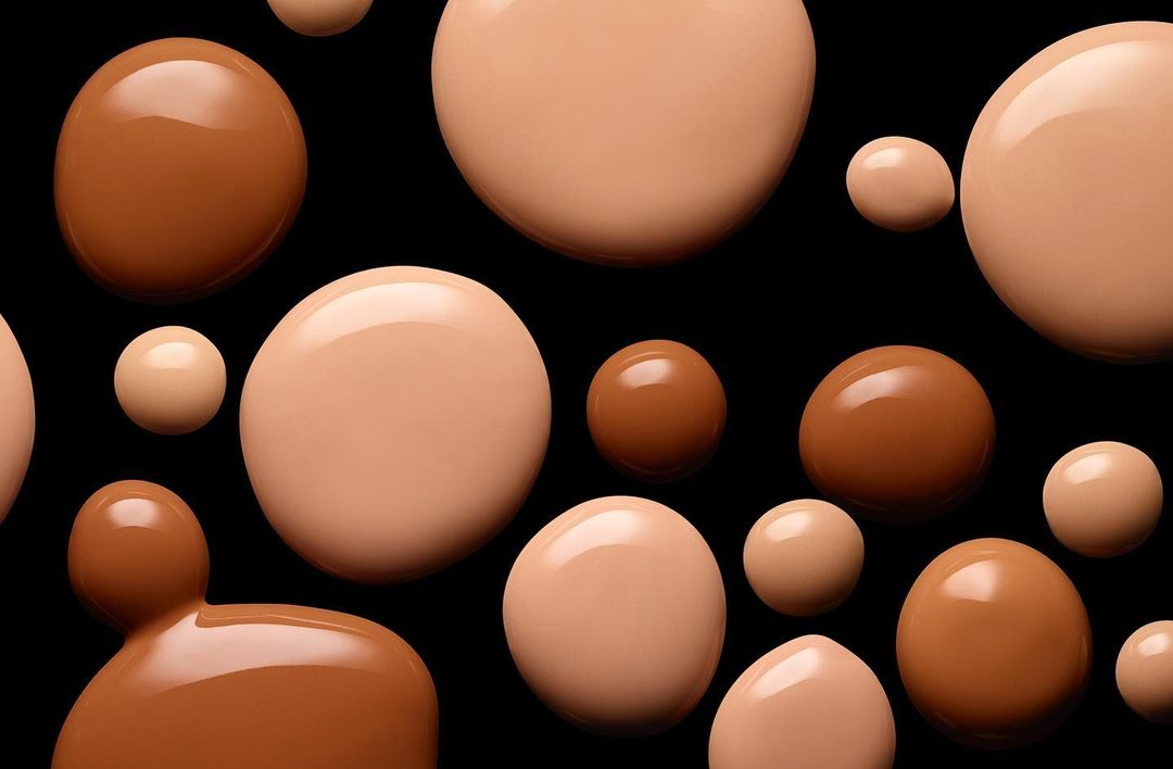Multiple drops of beige and brown foundation are arranged on a black background creating a textured, patterned beauty product shot.