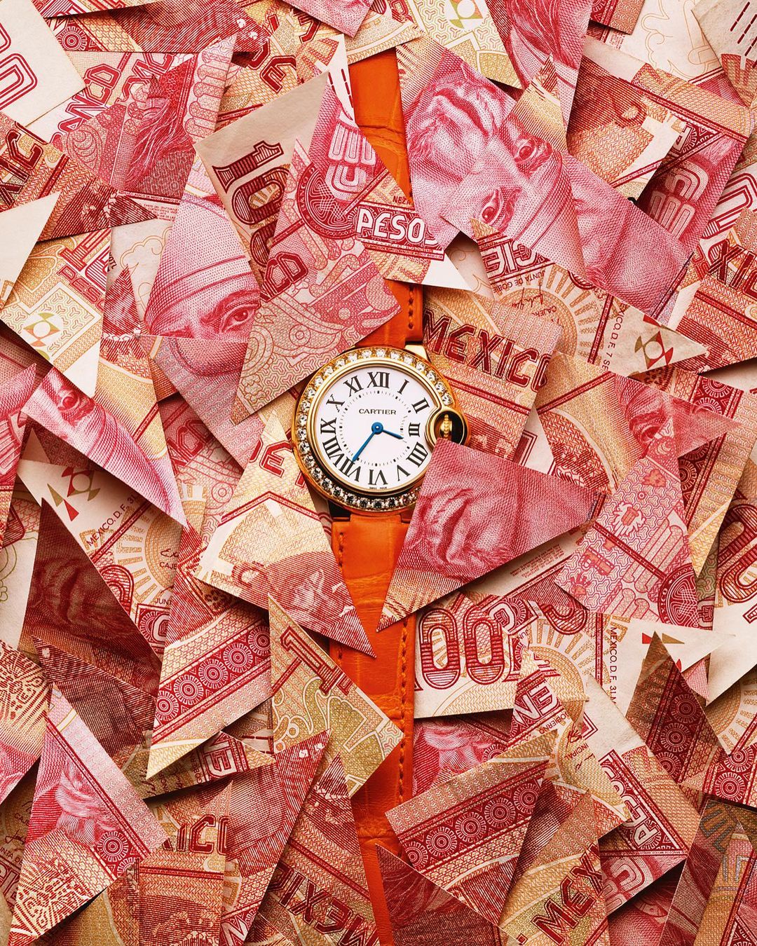 Wristwatch with a gold case and orange leather strap displayed atop a pile of cut-up Mexican currency, creating a visually striking composition.