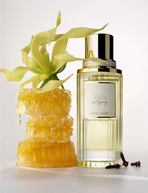 Close-up of Estee Lauder perfume bottle with gold cap next to a stack of honeycomb and yellow flowers on a white background.