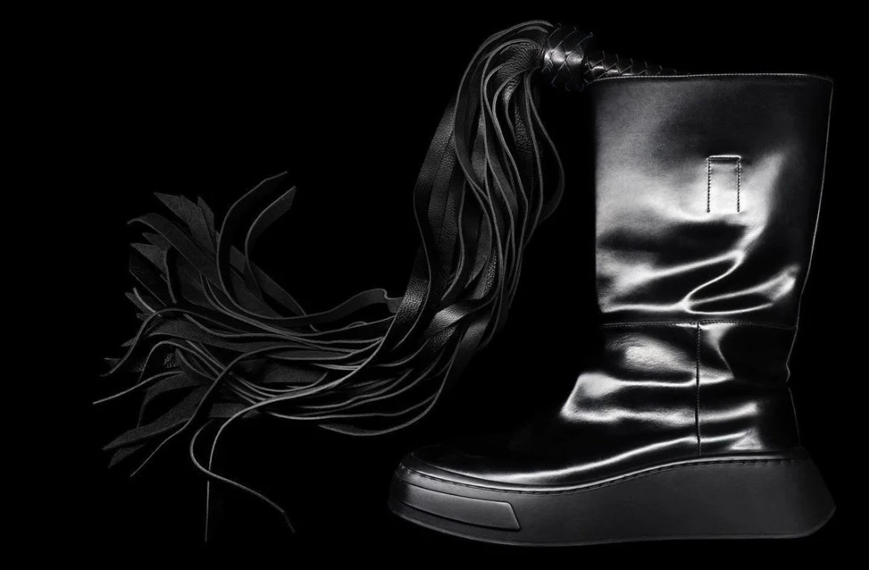 Fashionable black leather platform boot with an intricate, multi-strand leather tassel detail against a stark black background.