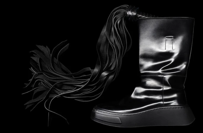 Fashionable black leather platform boot with an intricate, multi-strand leather tassel detail against a stark black background.