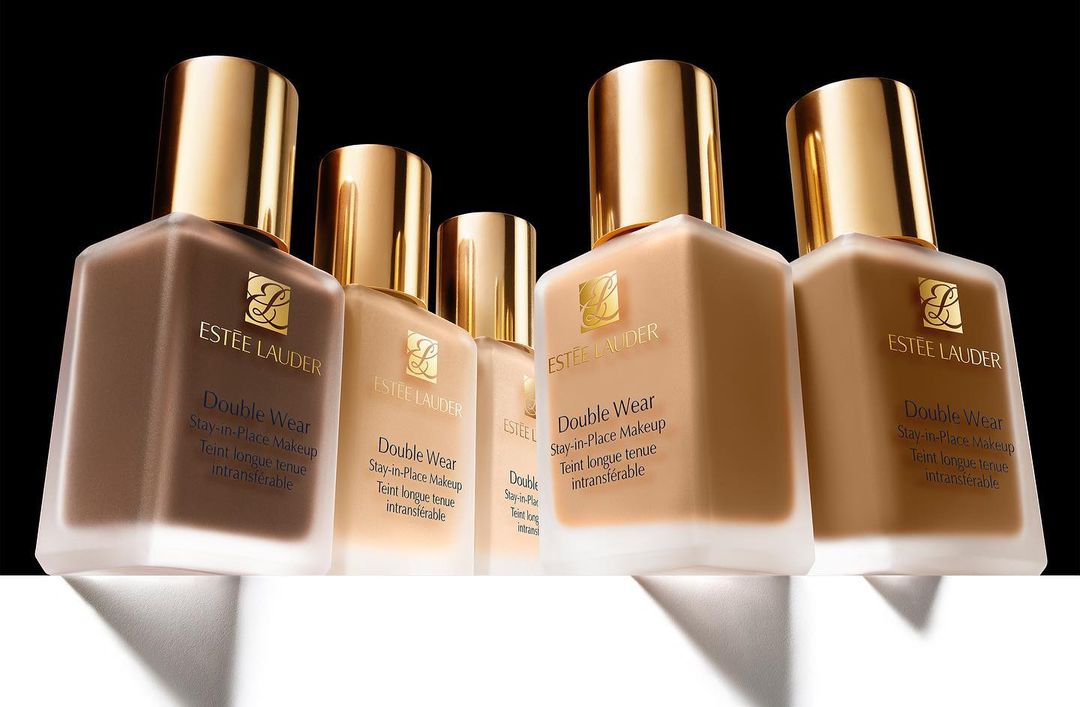 Multiple bottles of Estee Lauder Double Wear foundation in varying shades with gold lids on a white surface against a black background.