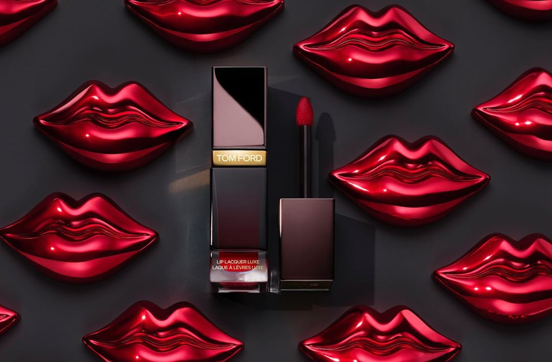 Close-up of Tom Ford red lip lacquer with applicator against a backdrop of glossy red lip-shaped decorations and a dark gray background.