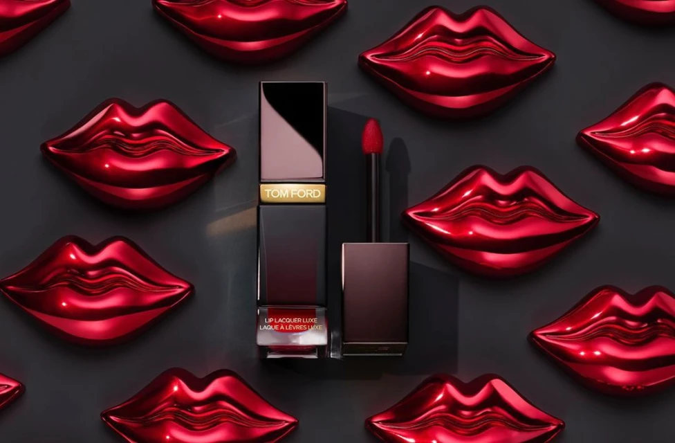 Close-up of Tom Ford red lip lacquer with applicator against a backdrop of glossy red lip-shaped decorations and a dark gray background.